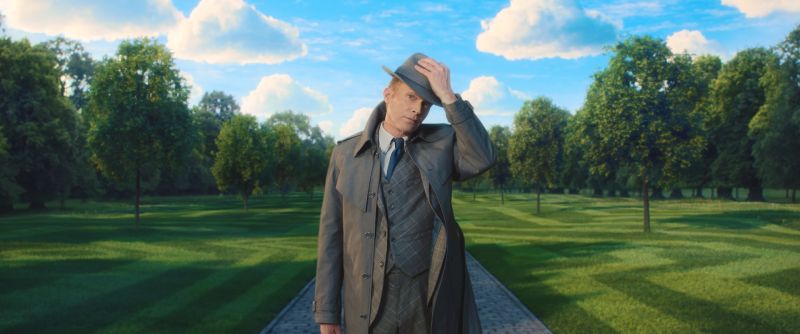 Paul Bettany luciendo sombrero de ala y gabardina gris en un amplio jardín verde bajo un cielo azul con nubes.