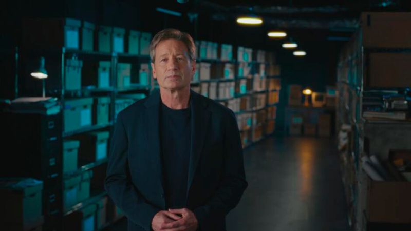 David Duchovny presenta la segunda temporada de Expedientes Desclasificados en HISTORY El actor y conductor David Duchovny parado de frente con las manos entrelazadas en primer plano. Viste un saco azul marino sobre una playera oscura. Se encuentra en una habitación iluminada de forma tenue con estanterías de metal repletas de cajas de archivo de cartón y expedientes. La iluminación ambiental resalta con tonos azules y cálidos al fondo.