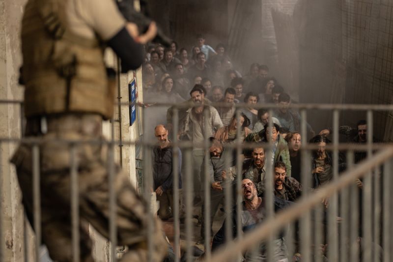 Fotograma de la película Apocalipsis Z: Nuevo Mundo. En el lado izquierdo, en primer plano de perfil y fuera de foco, se observa a un soldado armado sosteniendo un rifle de asalto. A la derecha, al fondo de un pasillo o rampa custodiada por una reja metálica, una multitud enfurecida de infectados se abalanza hacia la cámara con expresiones de desesperación y agresividad.