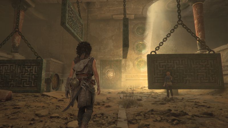 Exploración de ruinas en Resonance A Plague Tale Legacy Sophia caminando dentro de unas ruinas antiguas de piedra frente a grandes mecanismos colgantes grabados con laberintos.