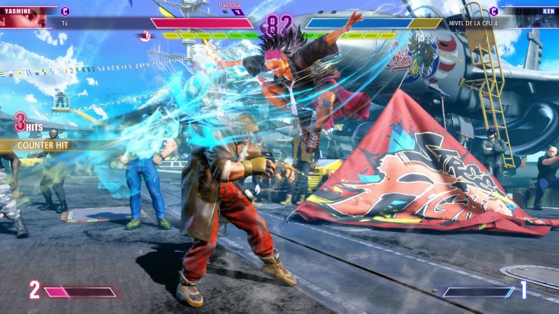Yasmine atacando a Ken desde el aire Captura de pantalla de pelea donde Yasmine salta por los aires ejecutando una patada descendente rodeada de ráfagas de viento contra Ken Masters.