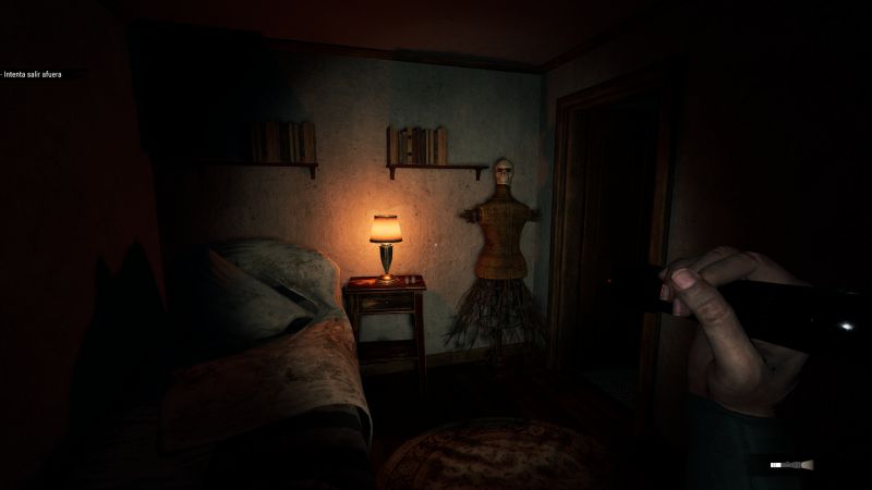 Habitación tenue con cama, lámpara y un maniquí junto a la puerta iluminado por una linterna.
