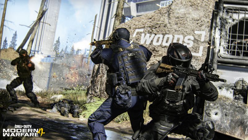 Captura de pantalla en tercera persona del multijugador de Call of Duty: Modern Warfare 4. En el centro, dos soldados con trajes tácticos oscuros y cascos de combate se cubren detrás de un montículo rocoso que lleva la palabra "WOODS" pintada en letras blancas. Uno de los soldados apunta hacia la izquierda con un rifle de asalto dorado, mientras el otro, en primer plano a la derecha, sostiene un rifle negro con ambas manos. A la izquierda, un tercer soldado avanza disparando su arma en medio de una explosión de tierra, y al fondo se aprecian árboles secos y una estructura de concreto gris bajo un cielo parcialmente despejado. El logo del juego aparece en la esquina inferior izquierda.