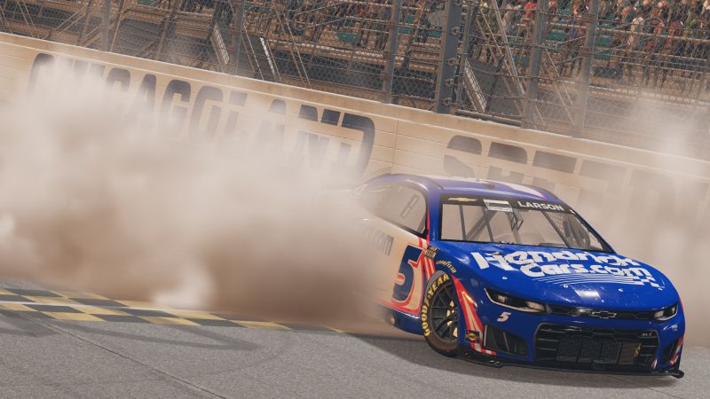 Captura de juego oficial de NASCAR 26 desarrollado por iRacing Studios Captura de pantalla directa del videojuego NASCAR 26 que muestra una dramática maniobra de derrape o burnout sobre el asfalto. En el centro de la toma destaca un auto de carreras Chevrolet Camaro en color azul brillante con detalles en blanco y rojo, marcado con el número 5 y el patrocinador HendrickCars.com. En la parte superior del parabrisas se observa el apellido del piloto Larson. Detrás del vehículo se genera una densa nube de humo blanco y polvo que cubre la barda de contención del circuito Chicagoland Speedway, con las gradas llenas de espectadores al fondo.