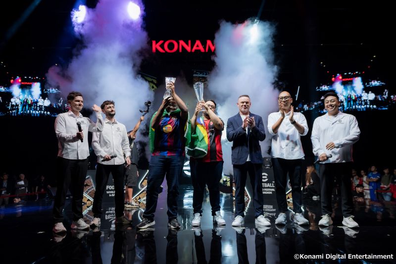 Escenario principal del eFootball World Festival en Bangkok lleno de luces dramáticas y humo blanco. En el centro del escenario, dos jugadores de esports vistiendo camisetas deportivas de fútbol y banderas de Brasil sobre los hombros levantan trofeos de cristal transparente sobre sus cabezas. A sus lados, organizadores con micrófonos, ejecutivos y la exfigura del fútbol Wayne Rooney vestido con saco oscuro y playera blanca aplauden el momento de la premiación. Al fondo se observa la pantalla gigante con el logotipo rojo de KONAMI.