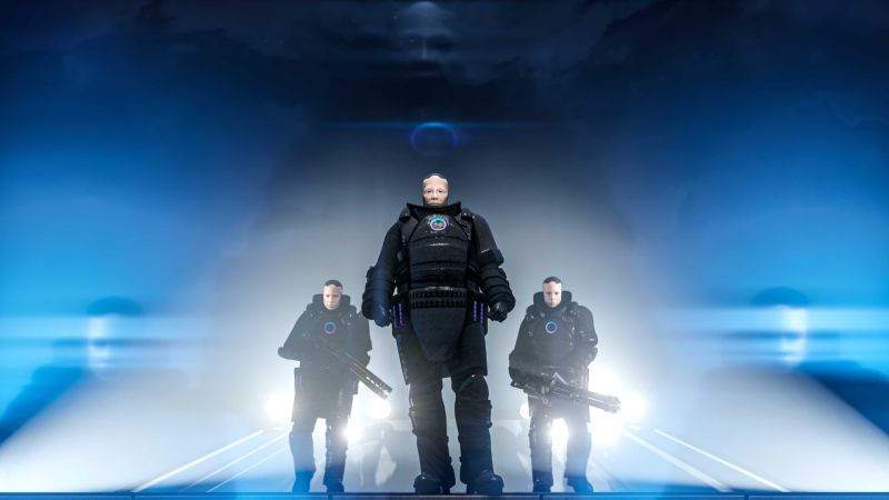 Tres personajes con armaduras futuristas posan en un entorno iluminado por luces azules y humo, evocando una escena de acción.