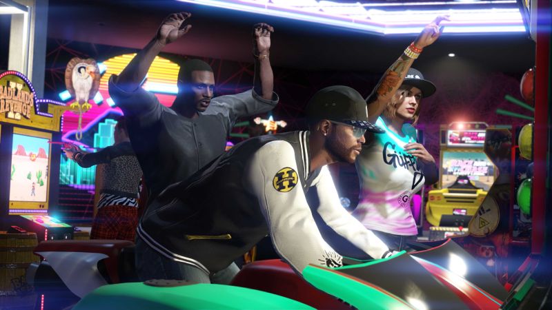 Captura de pantalla de un grupo de personajes dentro de un establecimiento de maquinitas recreativas en GTA Online. En primer plano, un hombre joven de espaldas con gorra negra y sudadera universitaria blanca y negra con un logotipo amarillo en la manga opera un simulador de carreras de motocicletas de color verde. Detrás de él, un hombre afroamericano con las manos levantadas en señal de festejo y una mujer con tatuajes en el brazo y gorra negra observan la partida con entusiasmo en un entorno iluminado por luces de neón moradas y pantallas de juegos arcade como Badlands Revenge II.