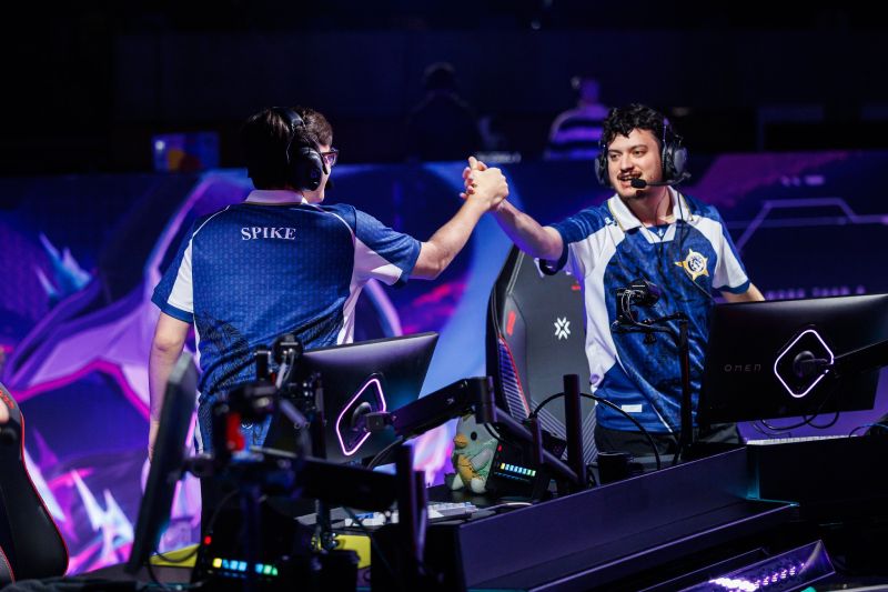 Dos jugadores de esports celebran en un escenario competitivo, vestidos con uniformes de equipo y rodeados de iluminación azul y púrpura.