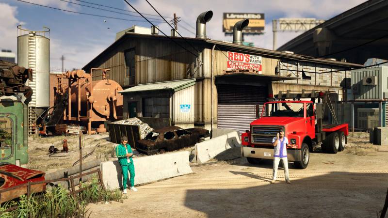 Imagen de dos personajes en un entorno industrial con un taller automotriz, maquinaria pesada y un camión de remolque, evocando la ambientación urbana característica de GTA Online.