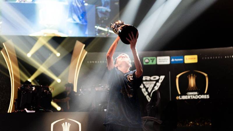 Fotografía de un jugador levantando un trofeo en un escenario iluminado, rodeado de pantallas y elementos visuales propios de un torneo profesional de esports.